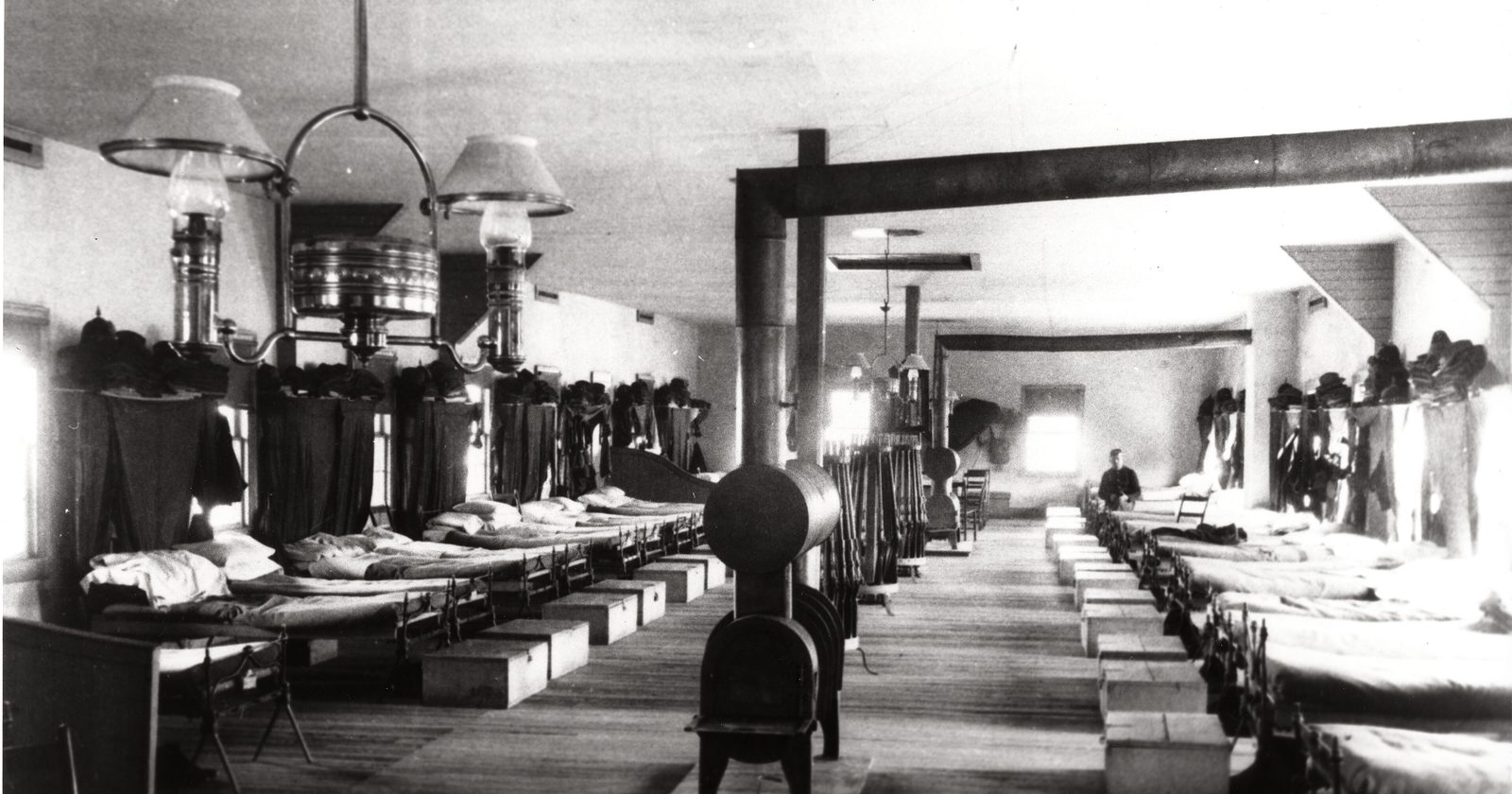 Archiwalna fotografia z lat 80. XIX wieku: długa sala sypialna z rzędami wąskich łóżek pod naftowymi lampami.